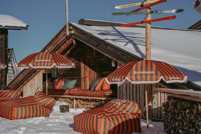 Schilala Aprés Ski Hütte von hinten mit Orange/ Grün gestreiften Schilala Kissen und Sonnenschirmen.