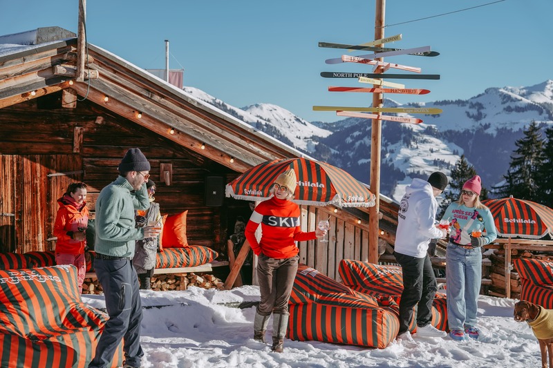 Mehrere Personen mit Getränken in der Hand auf der verschneiten Terasse der Schilala Aprés Ski Hütte in Kitzbühel.