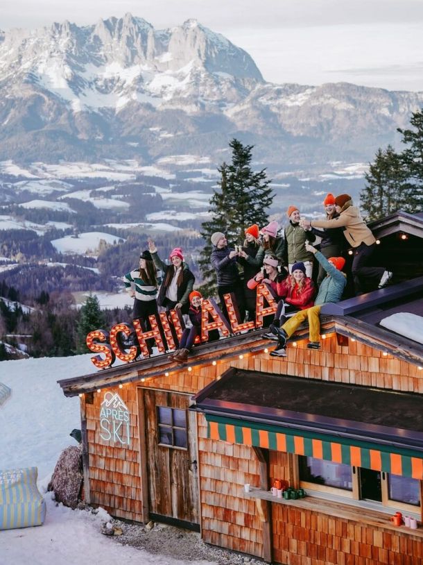 Ein Bild mit mehreren winterlich gekleideten Personen, die auf dem Dach der Schilala Hütte anstoßen. Blick auf die verschneite Berglandschaft.